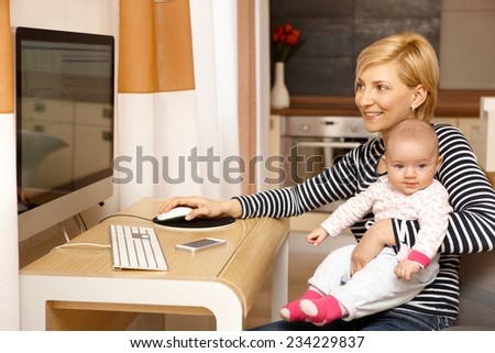 Young mother working on computer while holding baby girl on lap, smiling.