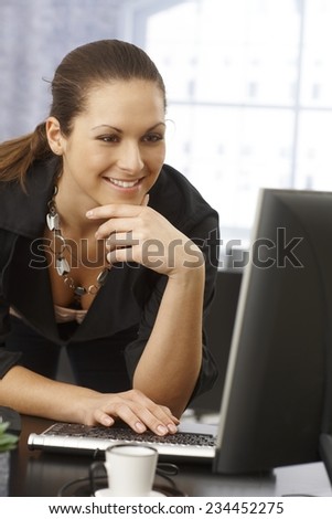 Happy businesswoman using computer in office, standing. leaning on desk.