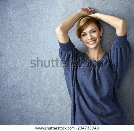 Happy young woman in blue jeans leaning against wall, smiling