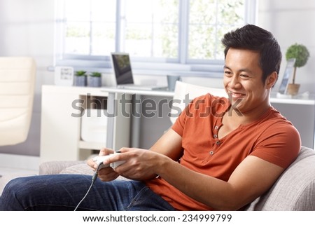 Young Asian man sitting in armchair at home, playing video game, smiing.