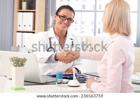 Happy female brunette doctor at medical office with patient, wearing glasses, stethoscope and lab coat. Shaking hands, smiling.