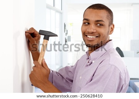 Happy young black man driving a nail to wall by hammer, smiling, looking away.