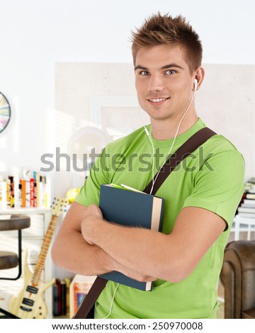 Portrait of happy male student smiling, holding books, listening to music through earbuds.