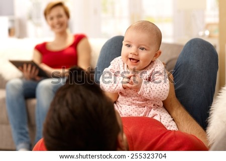 Baby girl and father playing on sofa, mother watching from background.