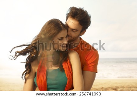 Attractive young casual caucasian brunette woman getting a kiss from boyfriend at beach. Romance, couple, smiling, embracing. Lens flare, sand and copyspace.
