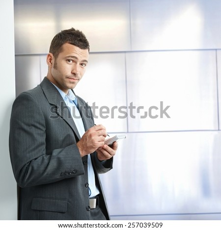 Annoyed young caucasian businessman in suit with tablet, standing in front of wall at business office. Looking at camera, copyspace.