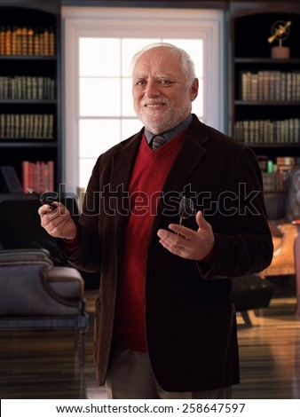 Happy caucasian senior man standing at retro home library room, wearing jacket, smiling, looking at camera, glasses and pen in hand, gesturing.