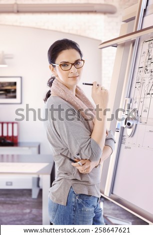 Casual female caucasian architect standing at drawing board and floor plan with pencil in hand at office. Wearing glasses, smiling, looking at camera.
