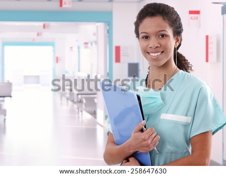Young happy afro american nurse standing at hospital ward with clipboard and pen in hand. Smiling, looking at camera.