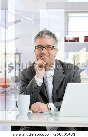 Happy mature businessman thinking at business office desk with laptop computer and coffee mug. Smiling, sitting, hand under chin, looking in the distance, wearing glasses.