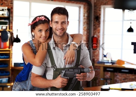 Happy young caucasian couple standing at home workshop, looking at camera, hugging, smiling. Power drill in hand.