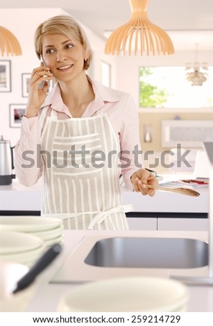 Blonde casual caucasian housewife in bright kitchen with mobile phone, indoors, standing, wooden spoon in hand, cooking, wearing apron.