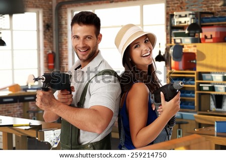 Happy young caucasian casual couple having fun at home workshop. Smiling, looking at camera, holding power drill in hand, standing. Handsome man, attractive female. Do it yourself.