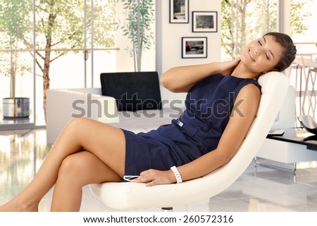 Attractive young brunette caucasian woman in blue dress resting in leather chair at trendy home. Smiling, caressing herself, eyes closed, relaxing.
