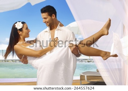 Young man holding beautiful bride in arms after dream wedding on tropical island, both smiling happy.