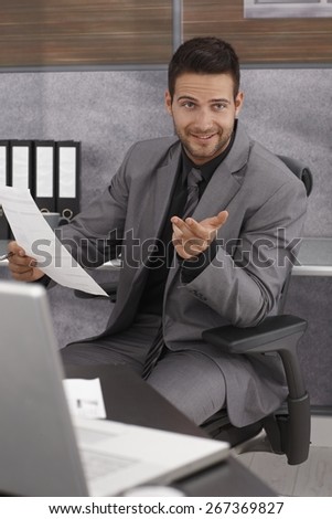 Office portrait of smiling young businessman sitting at desk with paper in hand, talking to someone.