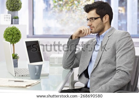 Young businessman sitting at desk, working with laptop computer, looking at screen.