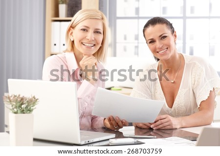 Happy casual blonde and brunette businesswomen at office working in front of laptop computer, checking business report papers, smiling, looking at camera.