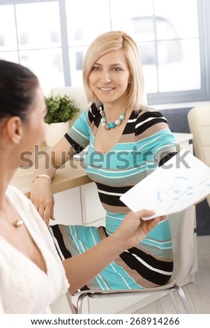 Blonde happy casual businesswoman working with colleague at office, sitting at desk, smiling, handing over paperwork.