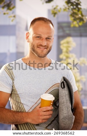 Closeup portrait of handsome young man holding bag and coffee in hands outdoors, smiling happy, looking at camera.
