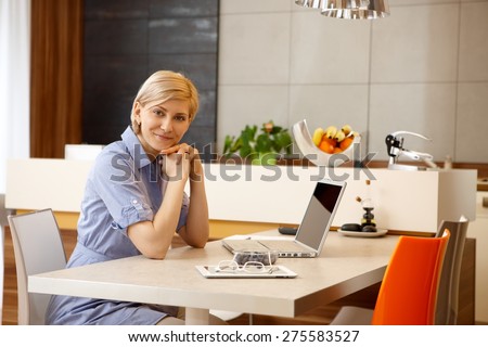 Blonde woman sitting at table at home, using laptop computer, smiling, looking at camera.