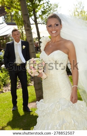 Beautiful woman in wedding gown smiling happy on wedding-day, wearing long veil, groom at background.
