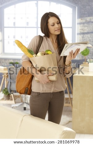 Young woman arriving at home, checking mail, holding shopping bag.