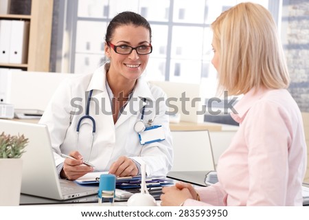 Happy female brunette doctor at medical office with patient, writing on clipboard, wearing glasses, stethoscope and lab coat.