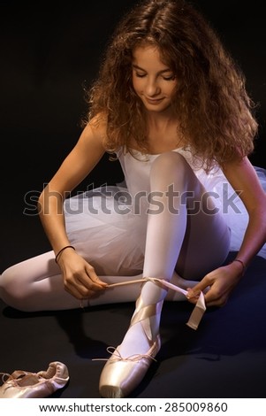 Pretty young ballet student tying lace on ballet shoe, sitting on floor.