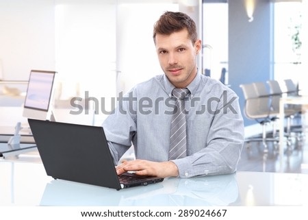 Young caucasian businessman in shirt and tie sitting at office desk using laptop looking up at camera.