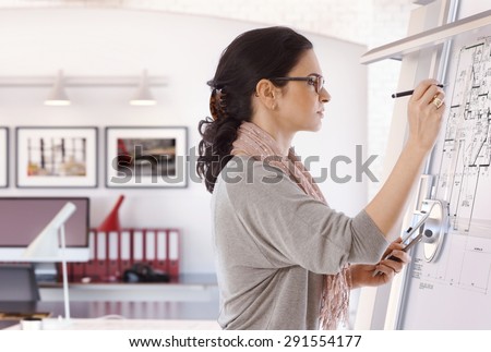 Focused casual caucasian female architect working at drawing board with pen in hand. Wearing glasses, at office. Floor plan, busy, concentration, unsmiling.