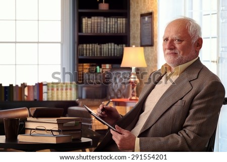 Senior caucasian man sitting in chair, wearing suit at home library, books on table, tablet computer in hand.