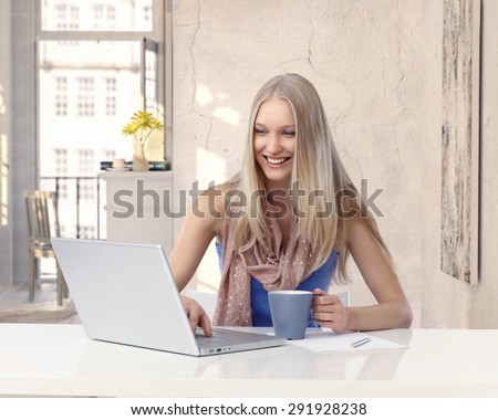 Happy young blonde casual caucasian woman working with laptop computer at retro home. Sitting at table with mug in hand doing business online, laughing, looking at screen.