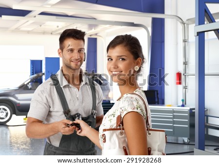 Happy attractive caucasian young lady at auto repair shop, getting car keys back from handsome dirty male mechanic. Smiling standing looking at camera.