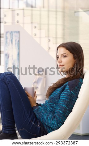 Happy young casual attractive caucasian woman daydreaming at home indoors, sitting in chair, cup in hand. Smiling, looking away,