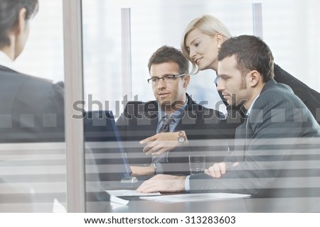 Focused caucasian business people meeting in boardroom behind glass. Sitting at table, wearing suit, looking at screen, pointing.