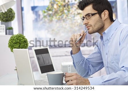 Young businessman sitting at desk, thinking, using laptop computer and tablet.