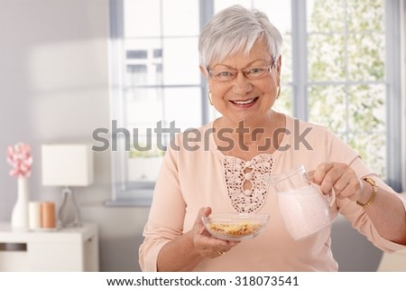 Happy elderly lady preparing breakfast cereal, pouring milk, looking at camera.
