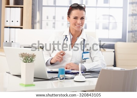 Happy brunette caucasian female doctor sitting at medical office desk in front of laptop computer, wearing glasses, stethoscope and lab coat.