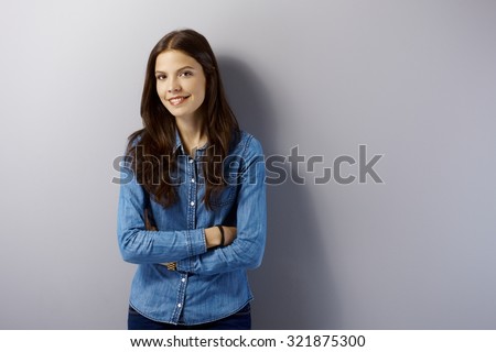Happy young woman standing arms crossed by grey wall, looking at camera.