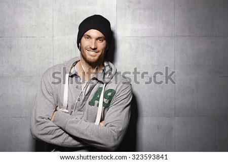 Happy urban man listening to music through earbuds, leaning against grey wall.