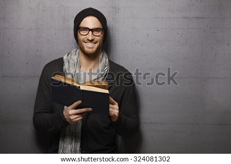 Happy urban style young man standing against grey wall, smiling, reading book, looking at camera.