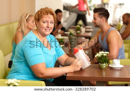 Young fat woman looking desperate, sitting at gym coffee table holding water bottle in hand.
