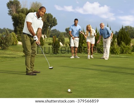 Male golfer putting in golf ball on the green, partners jittering.