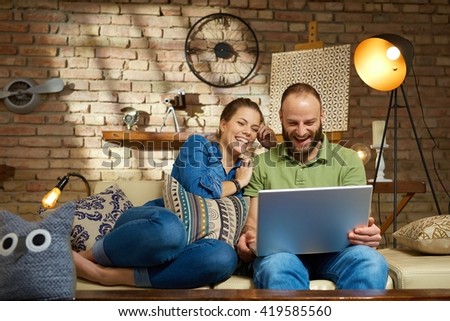 Happy couple using laptop computer at home, sitting on sofa, laughing.