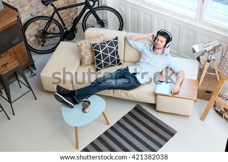 Young man lying on sofa at home, listening to music through headphones eyes closed.