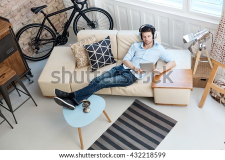 Young man relaxing on sofa, using tablet computer, listening to music at home.