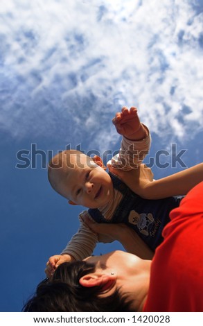 Baby and his mother are having outdoor fun together. The mother lifts him higher and higher and they are smiling a lot.
