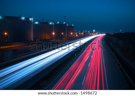 Heavy traffic on a highway. Due to the long exposure time the front and rear lamps of the cars are forming a white and a red snake of light. There are huge storage tanks on the left side.