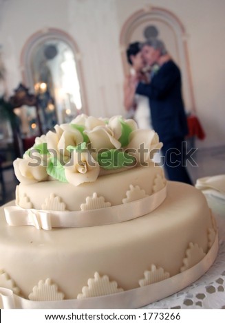 A wedding cake and the happy couple is dancing in the background. Focus is on the cake.